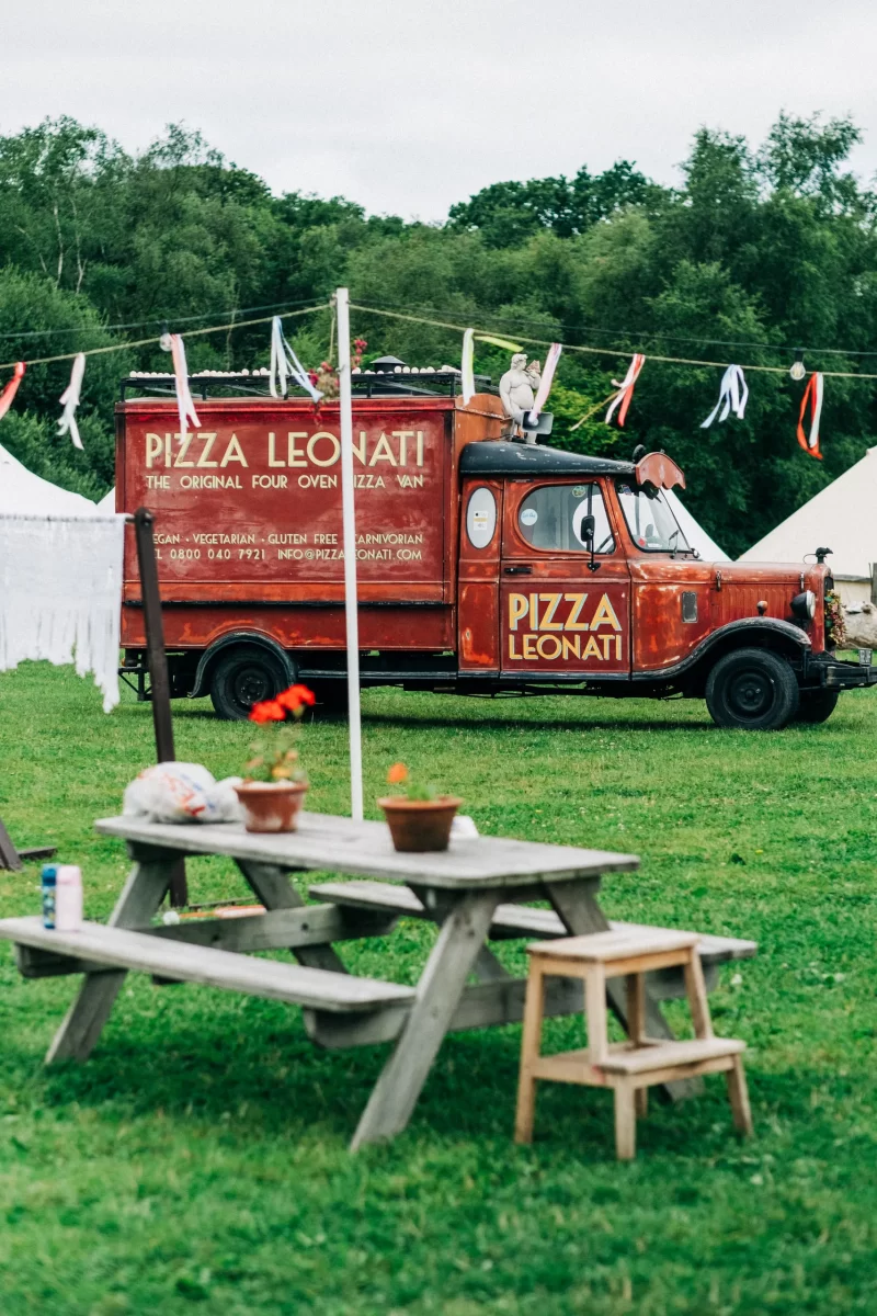 A vintage red pizza van labeled “Pizza Leonati” is parked on grass near white tents and trees—perfect for relaxed festival wedding vibes. A picnic table with flowerpots, a bench, and festive streamers offer charming garden festival wedding ideas at Frickley Lake near Battle