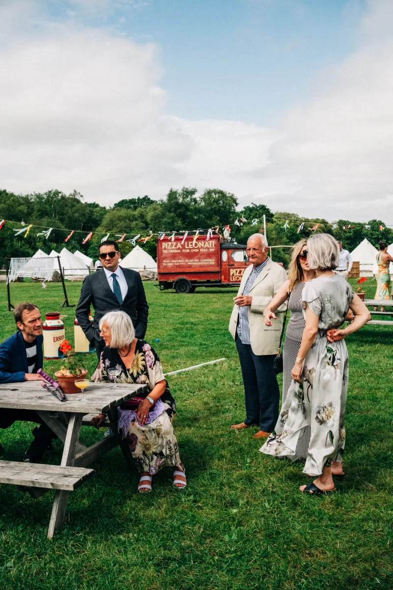 A group of people in formal attire gathers around a picnic table on a grassy field at a relaxed festival wedding, with tents, colorful bunting, and a red pizza food truck in the background—perfect for garden festival wedding ideas.