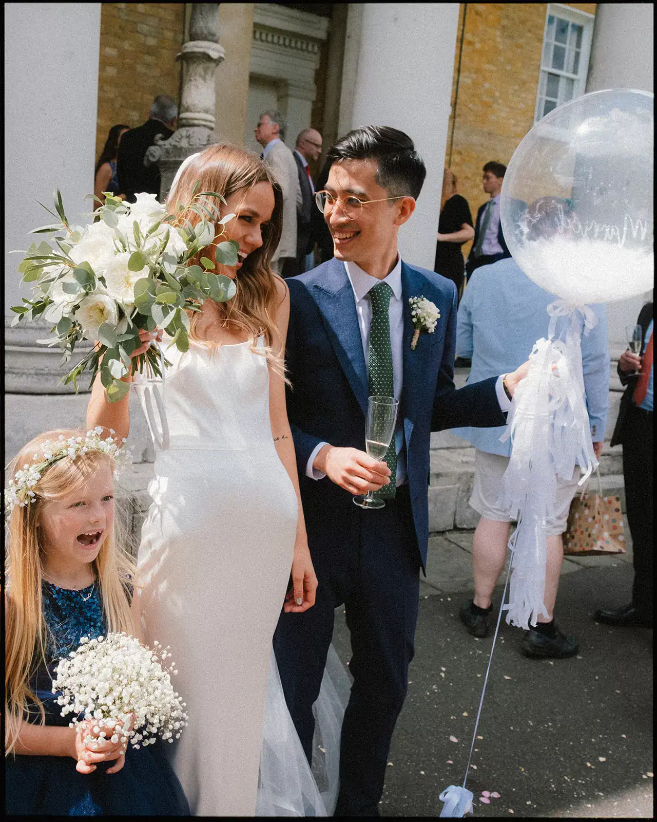 A bride in a white dress holds a bouquet and stands beside the groom in a navy suit holding a balloon at Clapton Country Club. A young flower girl in a navy dress with a floral crown smiles nearby as guests mingle in the background.