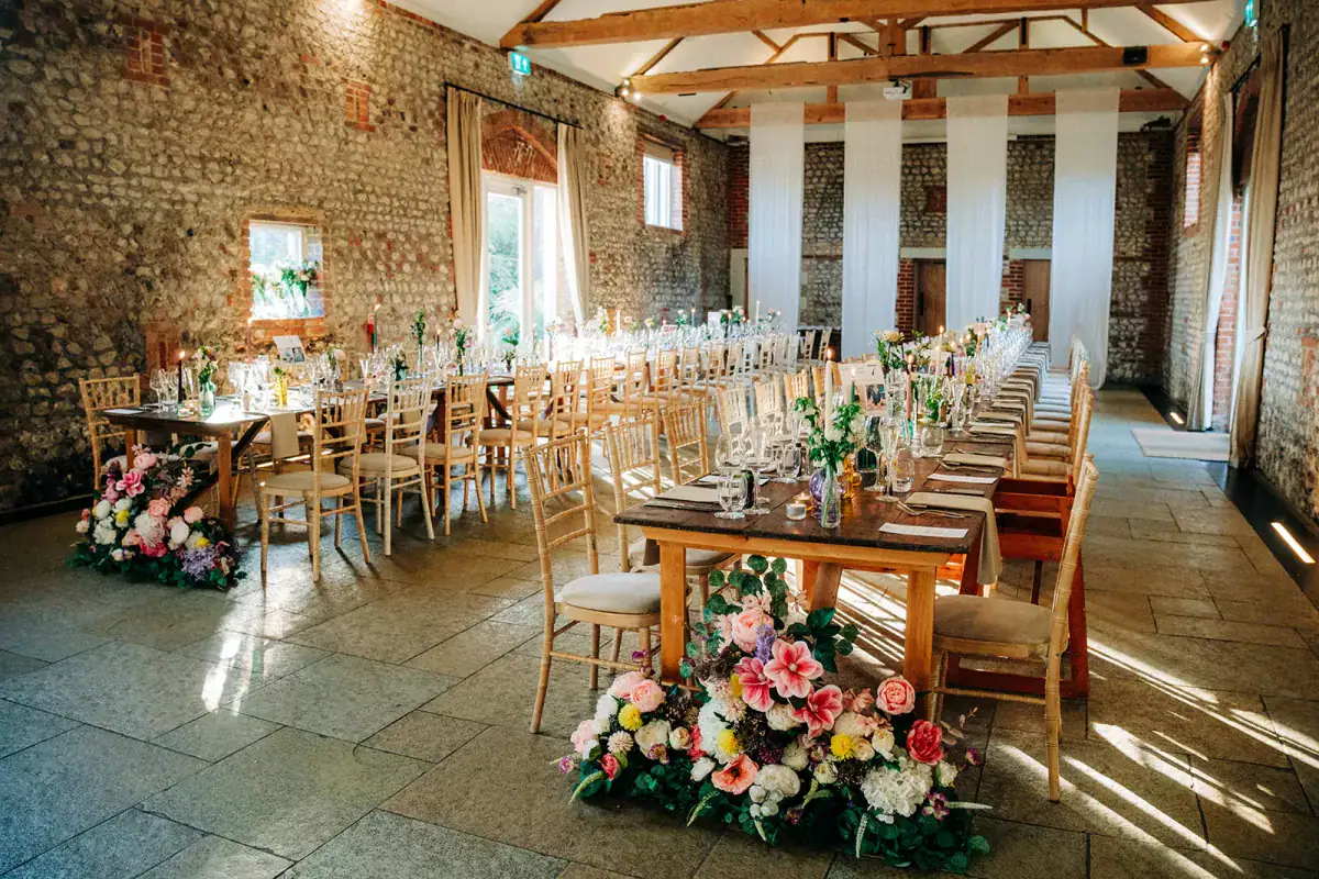 A rustic barn wedding venue near Chichester, Farbridge features stone walls, long wooden tables adorned with glassware, greenery, and vibrant flower arrangements at each end, with wooden chairs neatly arranged around the tables.