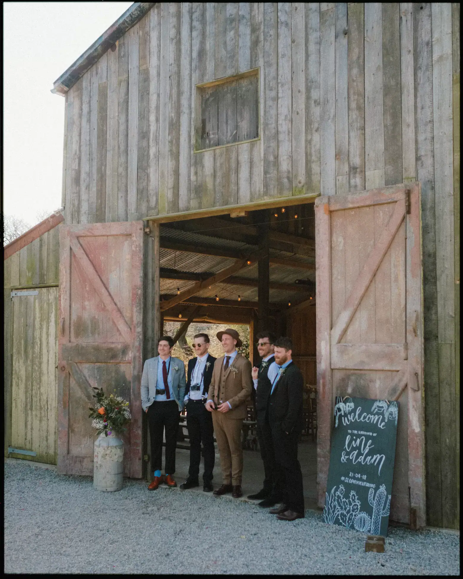 Five men in suits stand outside the rustic barn at Nancarrow Farm, Cornwall. Next to them, a chalkboard sign reads “Welcome Liv & Adam,” while a large floral arrangement enhances this charming rustic barn wedding scene.