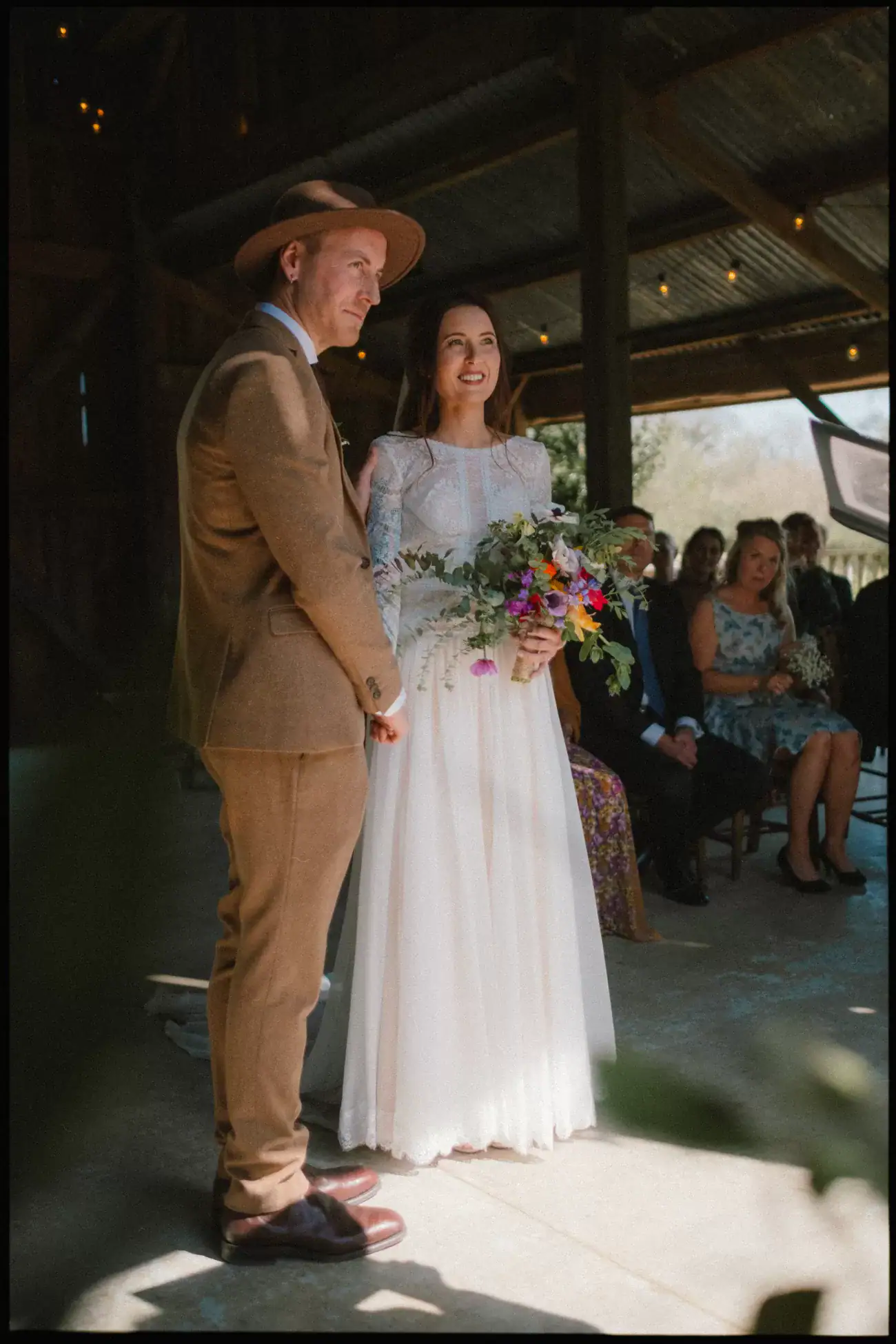 A bride in a white dress holding a colorful bouquet stands beside a groom in a brown suit and hat at an indoor wedding ceremony at Nancarrow Farm, Cornwall, with seated guests enjoying the charm of rustic barn weddings in the background.
