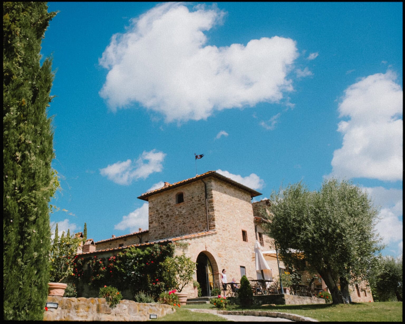 A rustic stone building with a red-tiled roof stands under a bright blue sky with fluffy clouds, surrounded by green trees and colorful flowers. Outdoor tables sit in a shaded garden area. Casa Cornacchi