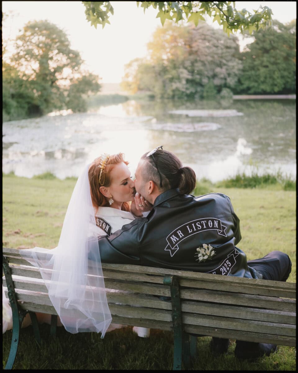 Newlyweds sharing a kiss by the lake at Preston Court during golden hour, wearing matching personalised jackets