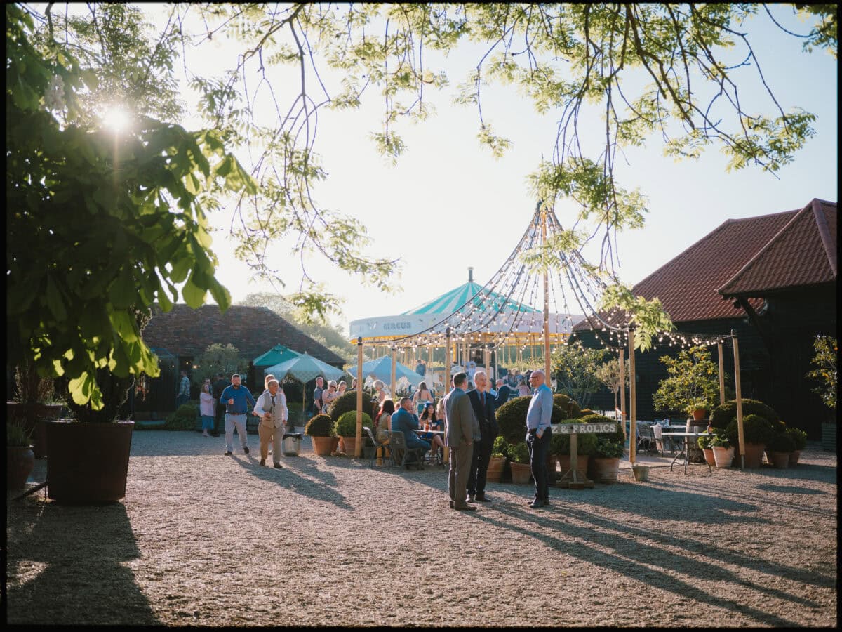 Courtyard at Preston Court wedding with guests mingling, carousel and festoon lights in evening sun.