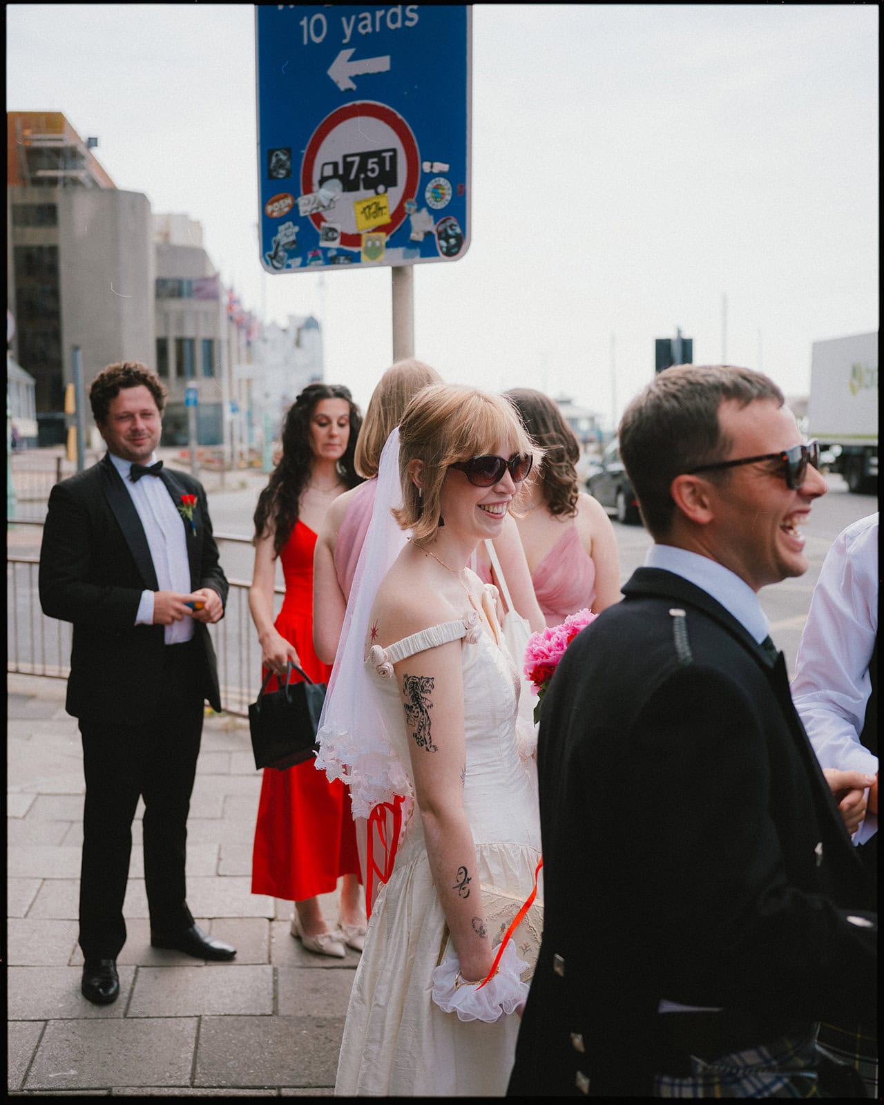 A bride in a white dress and sunglasses, holding pink flowers, stands smiling among friends in formal wear on Brighton seafront. Captured by an alternative wedding photographer Brighton, with a blue road sign and city buildings in the background.