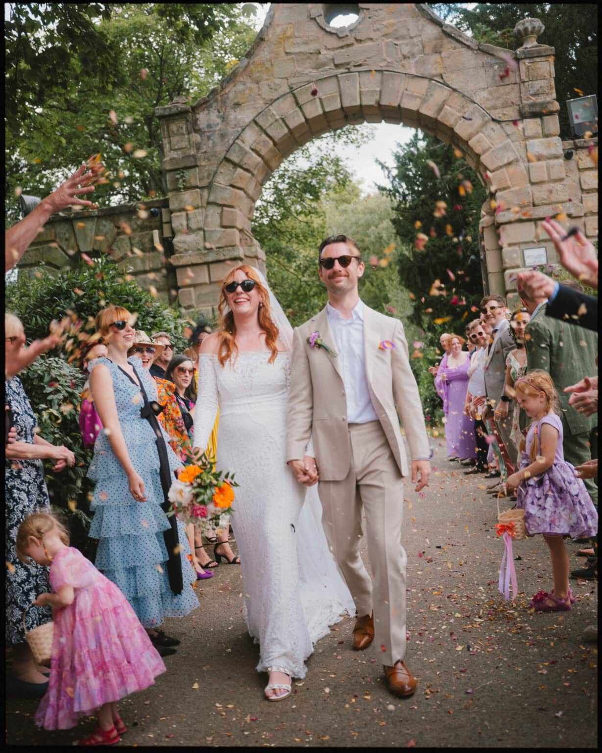 A bride and groom in sunglasses walk hand in hand under a stone archway, smiling as guests throw flower petals—a candid moment beautifully captured by an alternative wedding photographer London at this festive outdoor celebration.