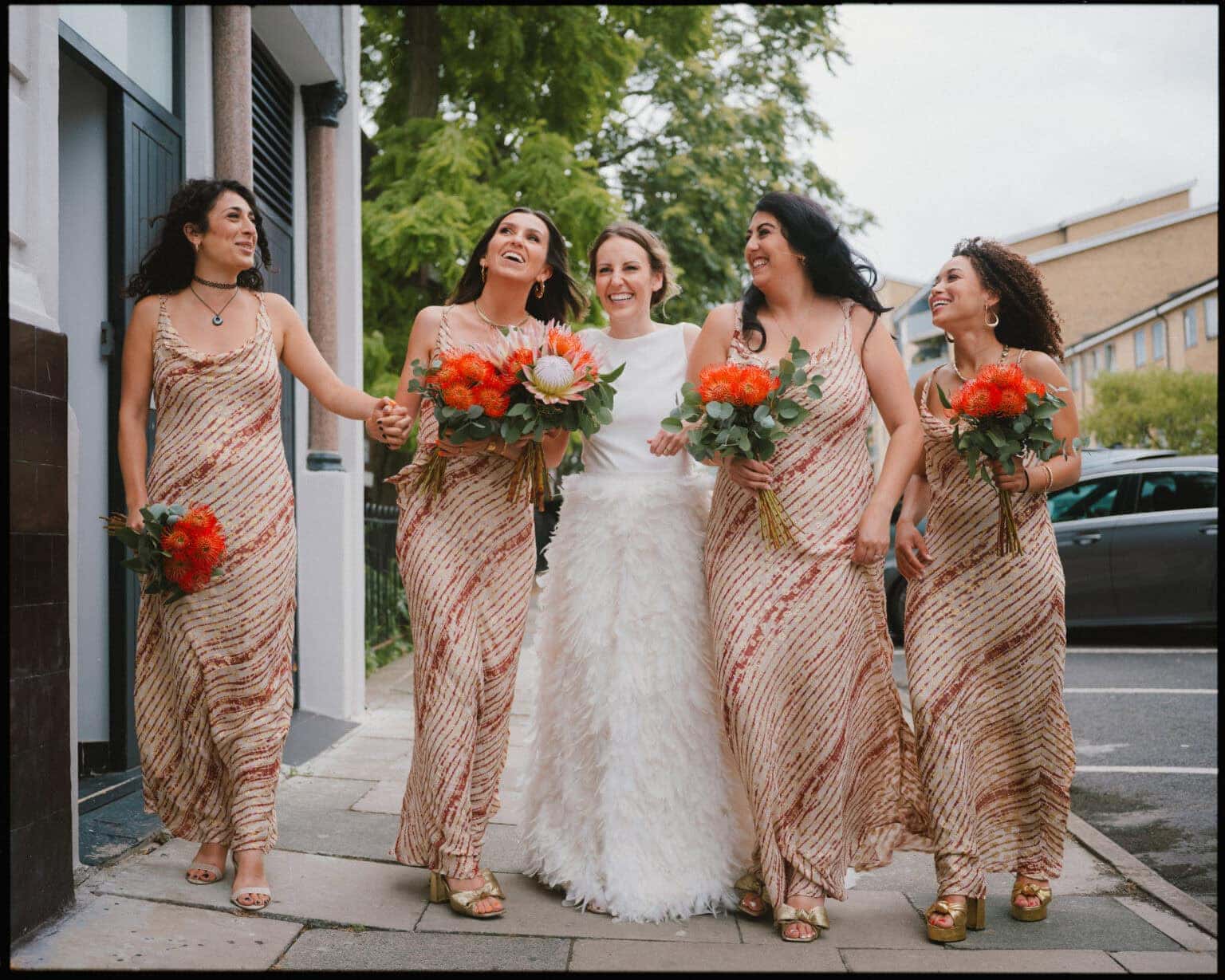A bride in a white dress stands on a sidewalk, smiling and holding hands with four bridesmaids in matching gold and pink dresses, each carrying bright orange bouquets—captured by an alternative wedding photographer London couples love.