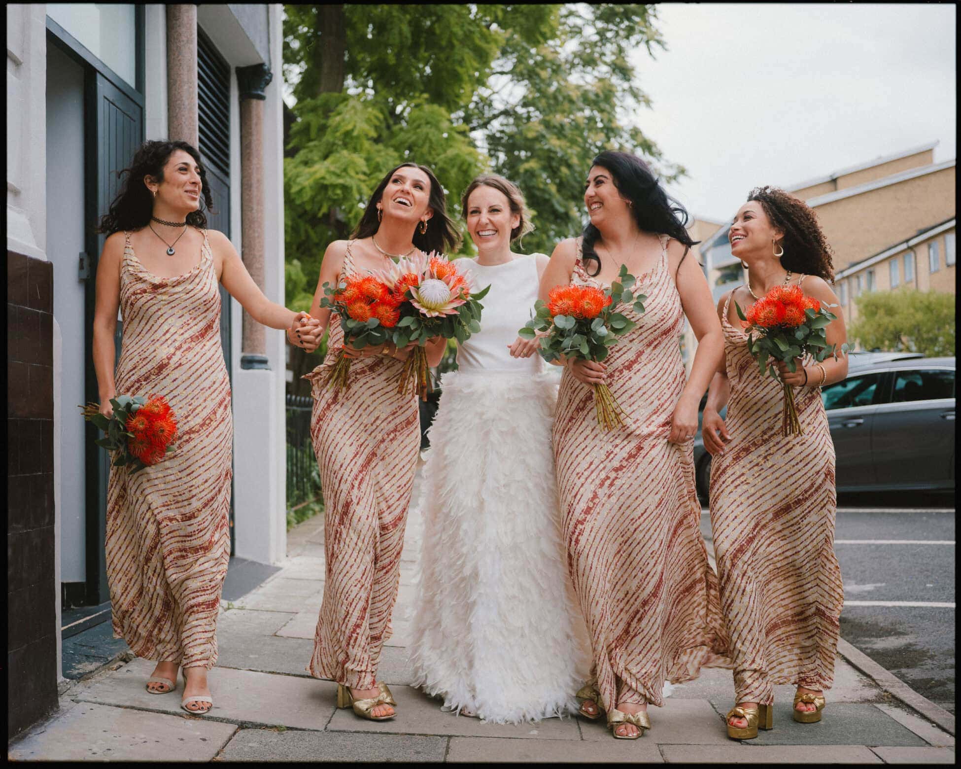A bride in a white dress stands on a sidewalk, smiling and holding hands with four bridesmaids in matching gold and pink dresses, each carrying bright orange bouquets—captured by an alternative wedding photographer London couples love.