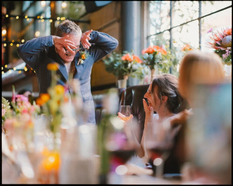 A man playfully makes a face with his hands at a lively, flower-filled indoor gathering, as a woman laughs and holds a drink. Other guests and drinks surround the decorated table—perfect for an alternative wedding photographer London moment.