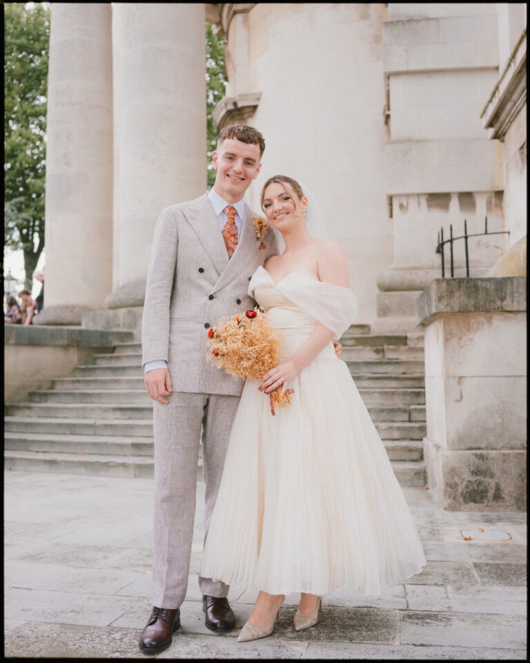 A bride and groom stand smiling together outdoors at their Greenwich Yacht Club wedding. The groom wears a light gray suit and tie, and the bride wears an off-the-shoulder white dress, holding a bouquet, with stone steps and columns in the background.