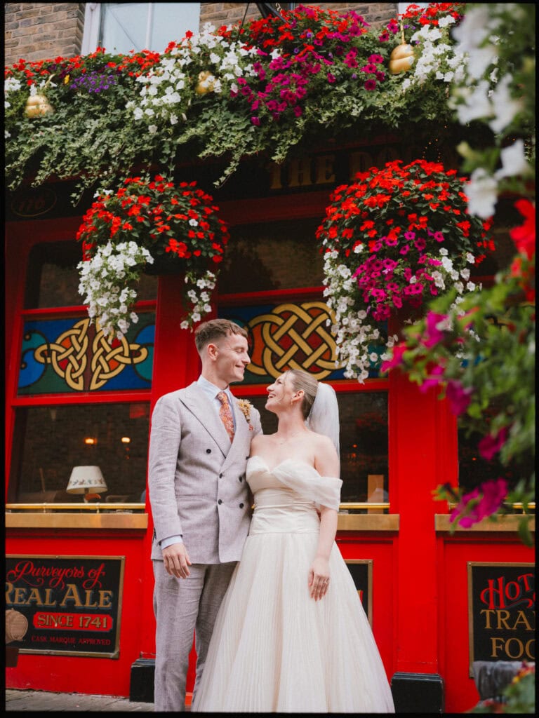 A bride and groom stand smiling at each other in front of a red pub adorned with lush hanging flowers. Captured by a Greenwich wedding photography expert, the bride’s off-shoulder gown and the groom’s grey suit shine on their special day.