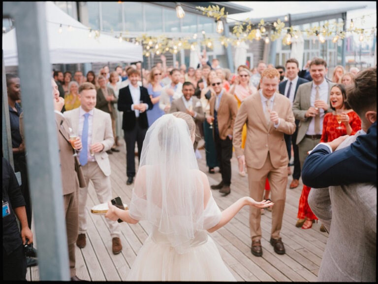A bride in a veil stands with her back to the camera facing a crowd of well-dressed guests at an outdoor London docklands wedding reception, with string lights overhead and people smiling and watching her.