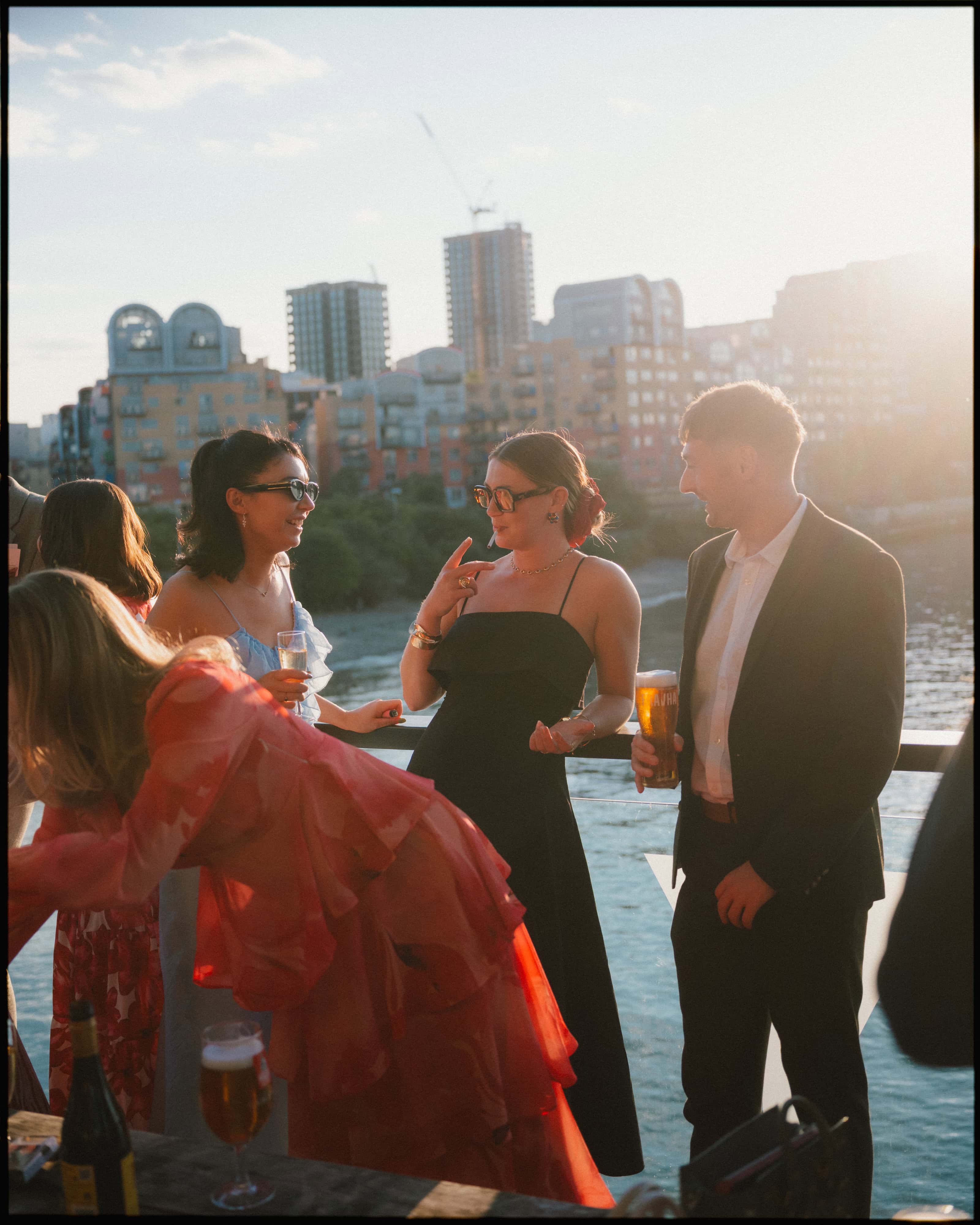 Guests chat with pints and wine on the Greenwich Yacht Club terrace in golden evening light, city skyline behind