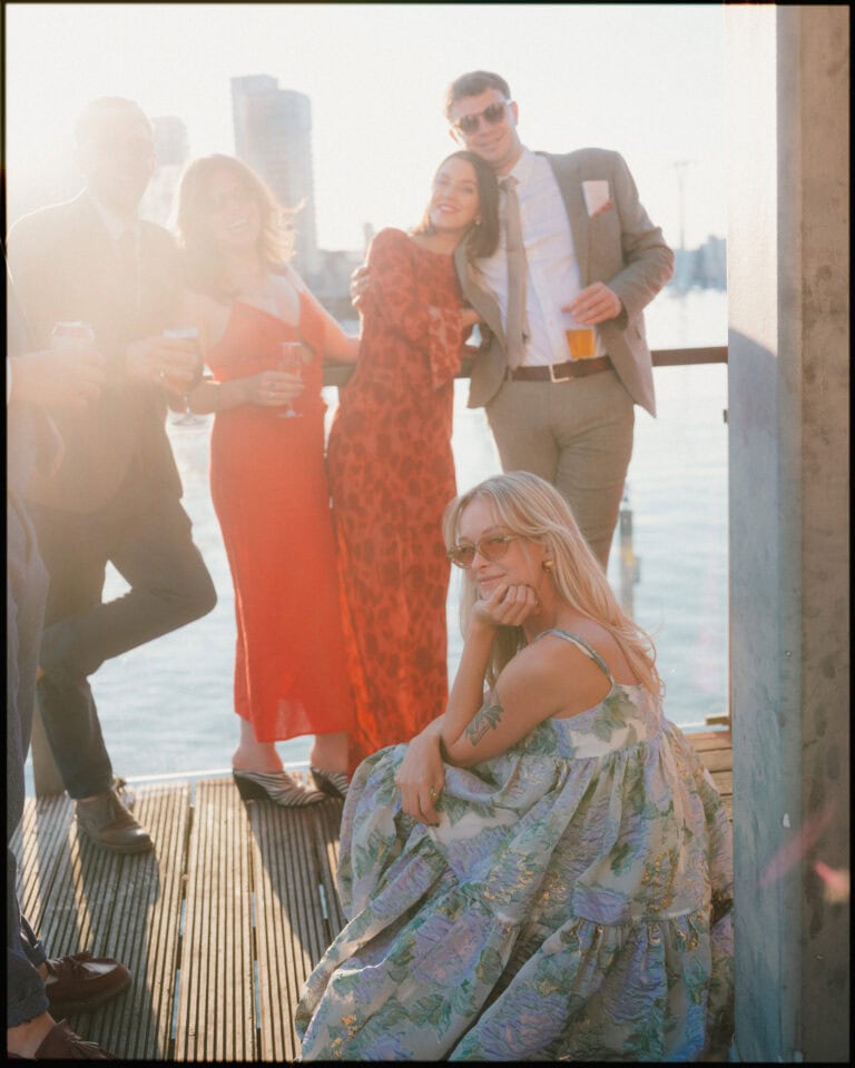 A group of four people in formal attire stand together on a sunny waterfront deck at a London docklands wedding, smiling, while a woman in a floral dress sits in front, posing and looking at the camera. City buildings and water are in the background.