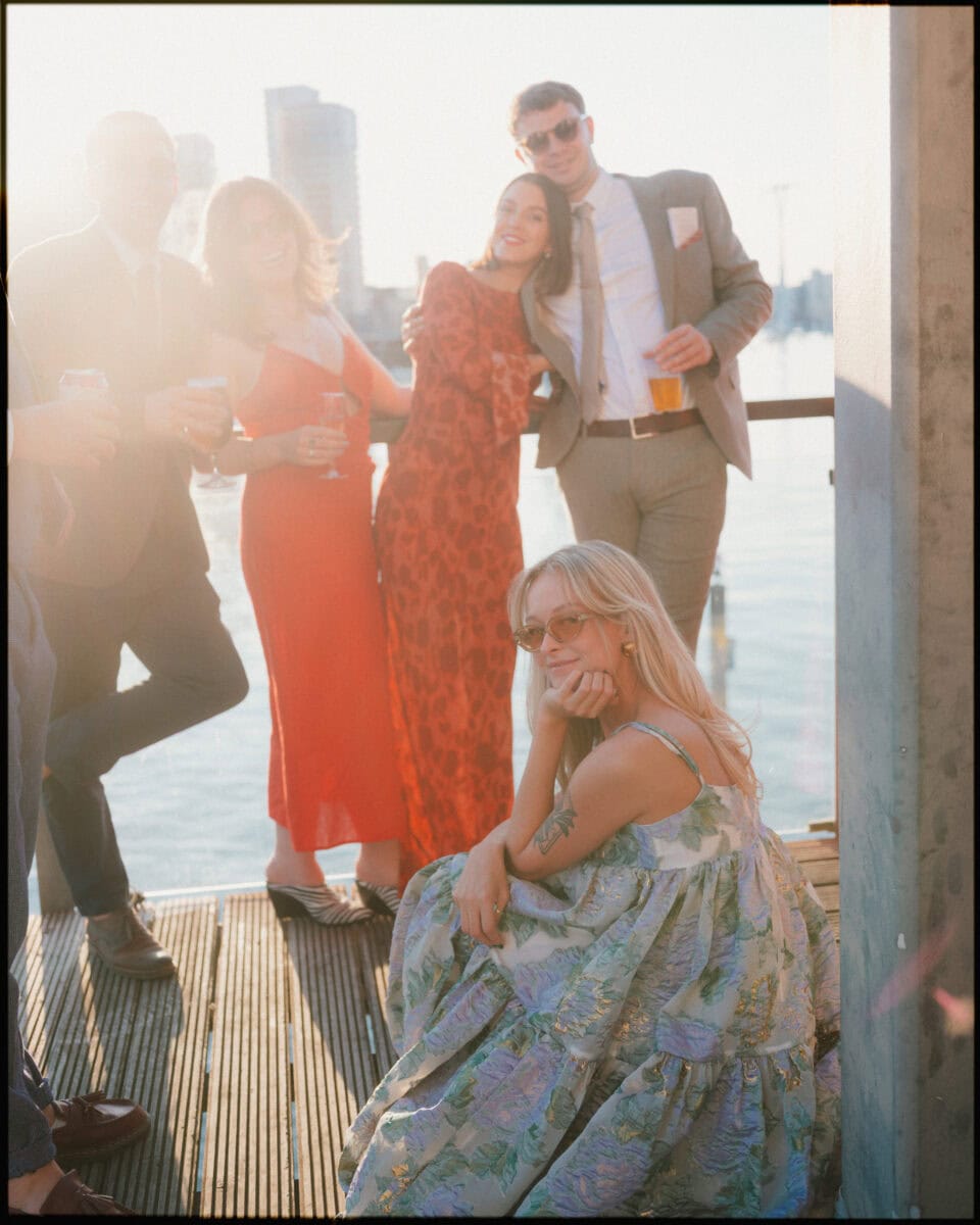 A group of four people in formal attire stand together on a sunny waterfront deck at a London docklands wedding, smiling, while a woman in a floral dress sits in front, posing and looking at the camera. City buildings and water are in the background.