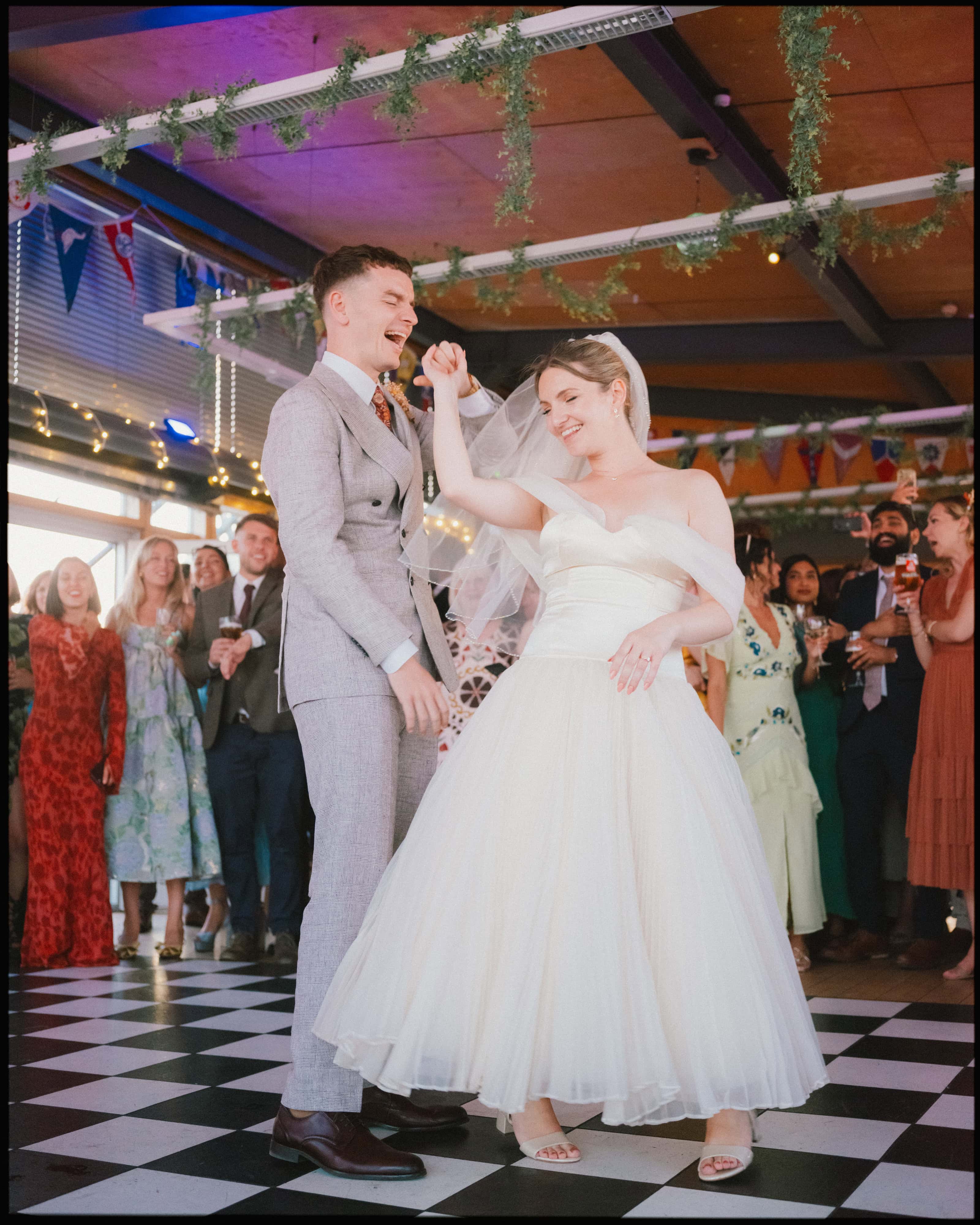 Groom in a grey suit grins as the bride in a tea-length gown dances on a chequered floor, guests cheering behind