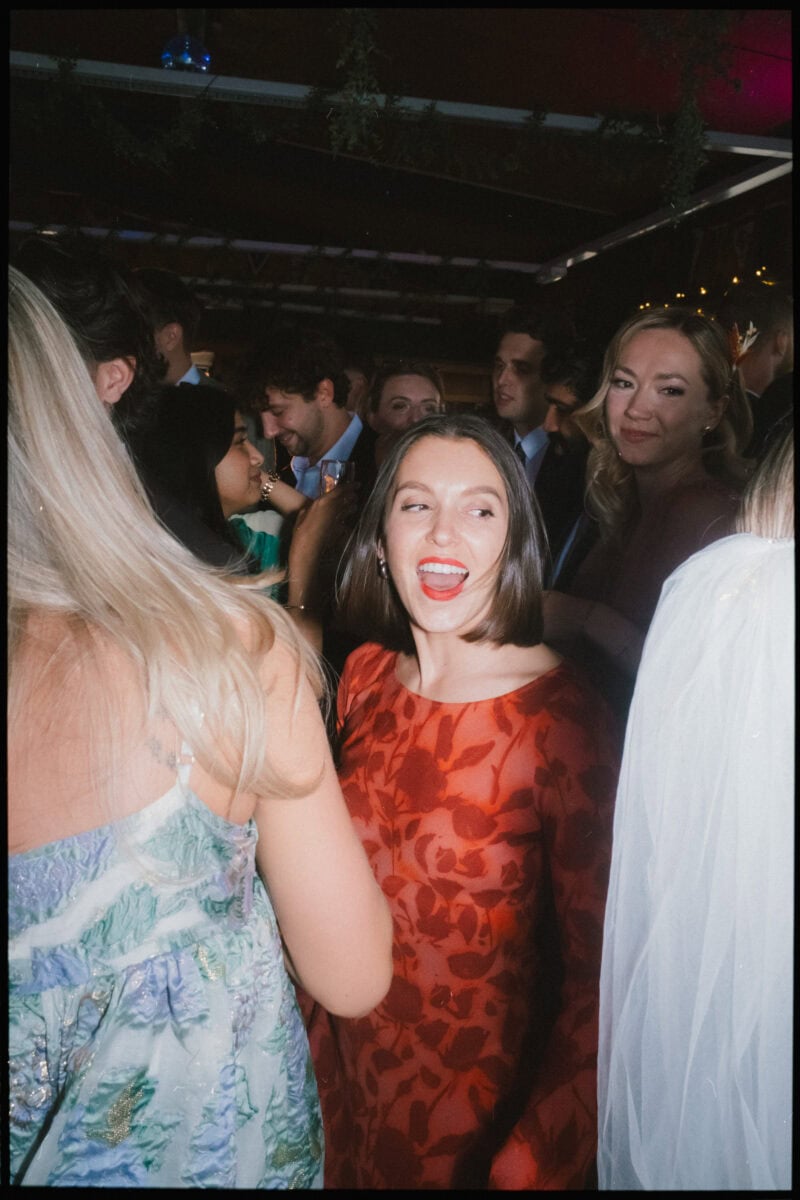 Woman in a red leaf-print dress laughs with her mouth wide open as guests crowd around her in a dim, beamed room