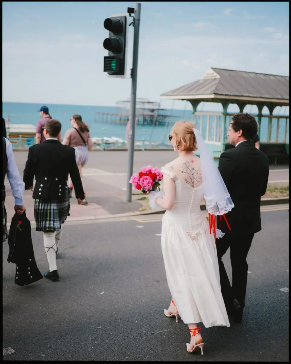 A bride in a white dress and veil holding pink flowers walks with a groom near the seaside. Captured in candid wedding photography, they cross a street by the pier, with a traffic light and others in the background.
