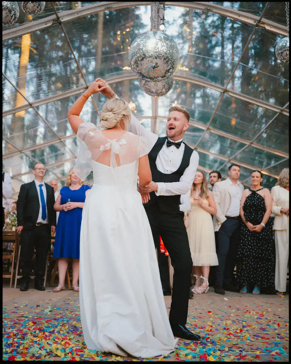 A bride and groom dance joyfully under disco balls in a glass-ceiling venue, surrounded by cheering guests and vibrant confetti—capturing the spirit of colourful editorial weddings with candid wedding photography.