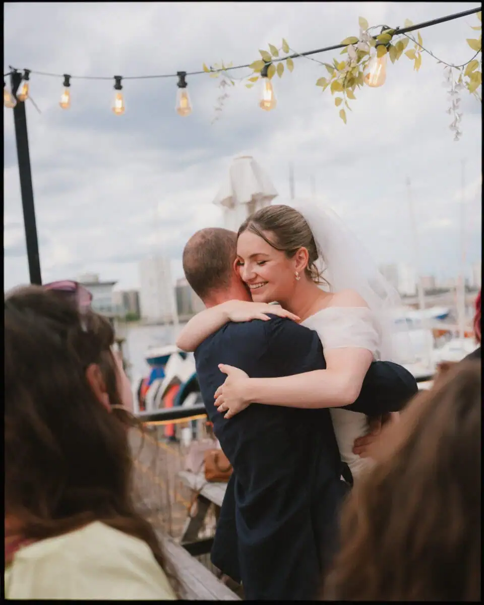 A bride in a white dress and veil hugs a man in a suit at an outdoor reception, with string lights above and boats behind—capturing the joy of the moment in candid wedding photography as guests watch the celebration unfold.