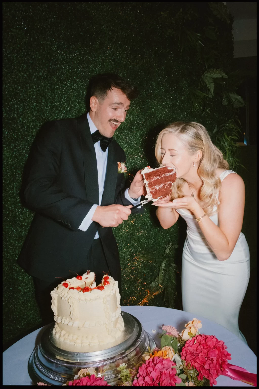 A man in a tuxedo playfully offers cake to a woman in a white dress, both laughing beside a decorated cake, framed by lush greenery—the perfect moment captured through documentary wedding photography.