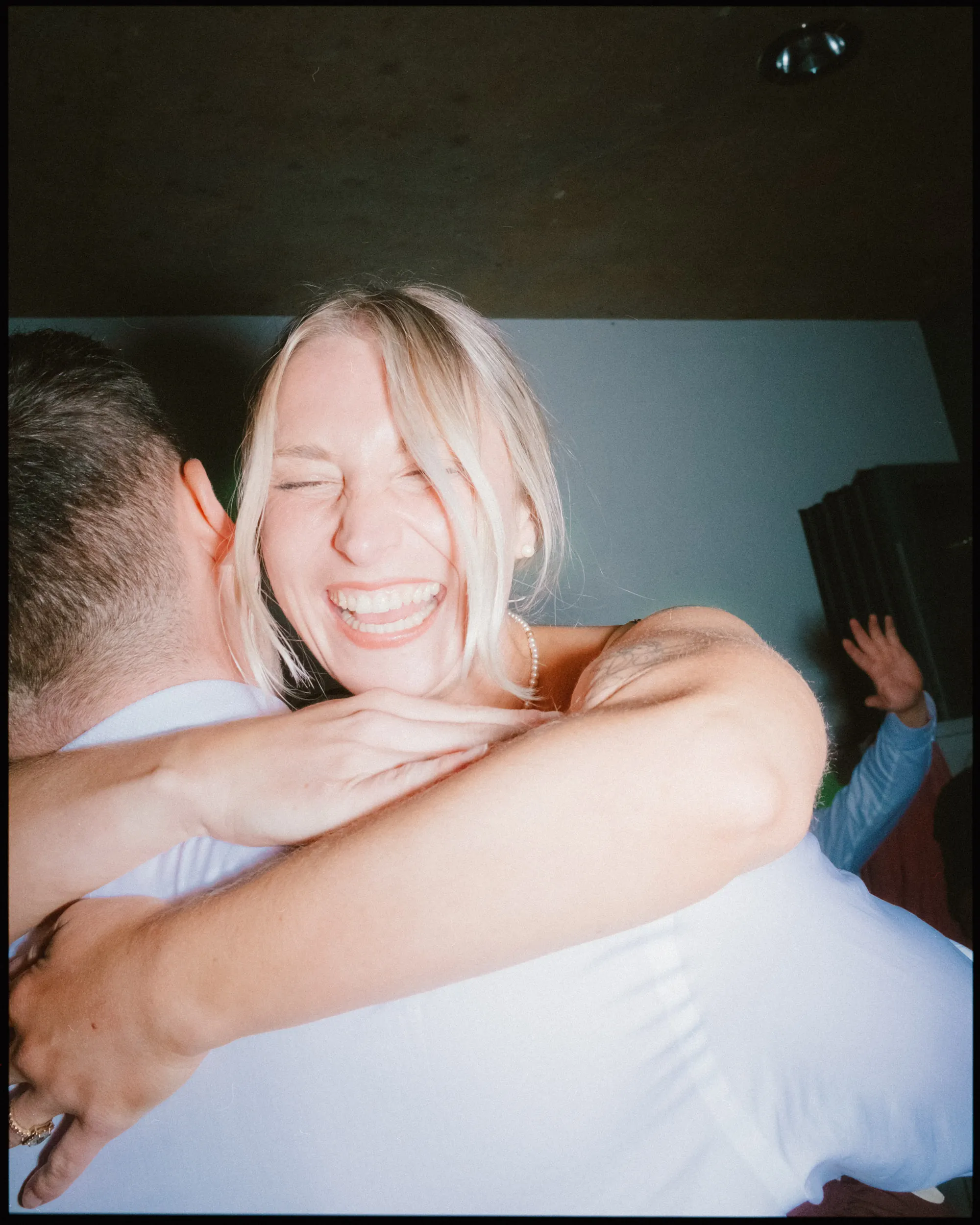 A woman with blonde hair smiles widely and hugs another person tightly, her eyes closed in joy, as another’s hand is raised in the background at what appears to be a party—capturing a perfect moment of documentary wedding photography.