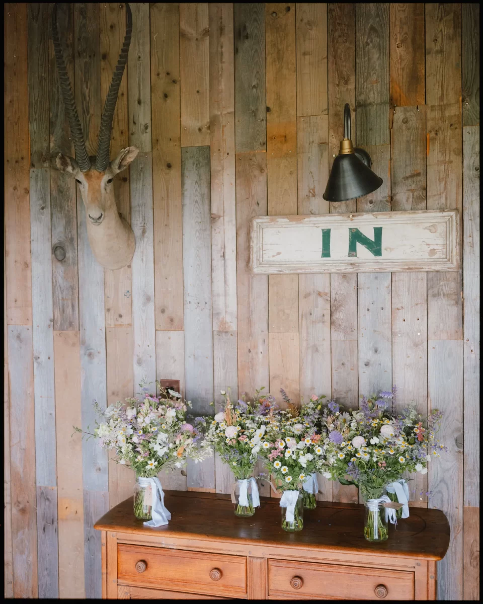 A wooden dresser holds six jars of wildflowers with white ribbons, set against a rustic wooden wall at a Montague Farm Wedding, with a mounted antelope head, a lamp, and a white sign reading IN—perfect for festival-style Sussex weddings.