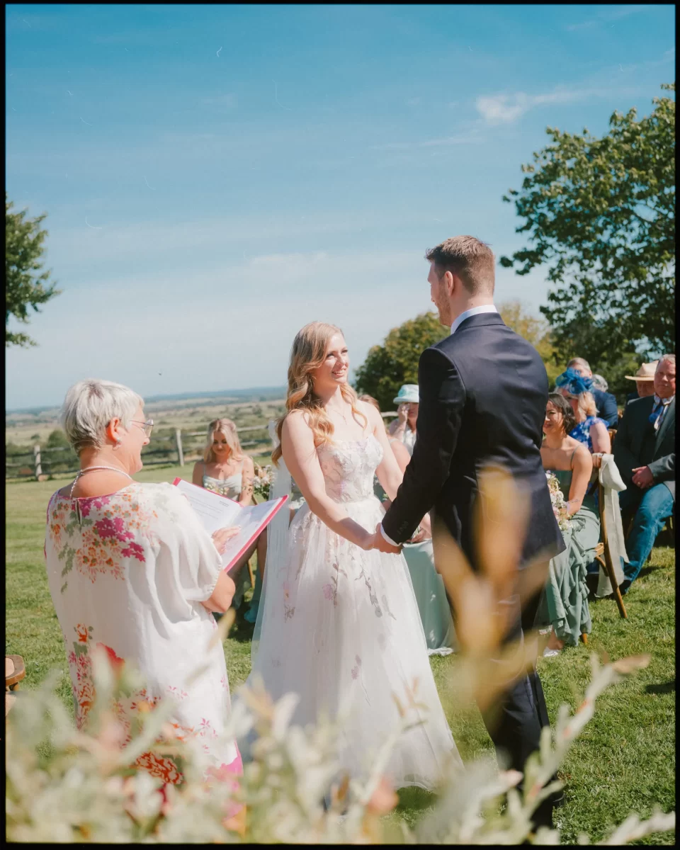 A bride and groom stand holding hands during a Montague Farm wedding, facing each other as a celebrant reads from a book. Guests are seated in the background, enjoying a sunny day and greenery at this festival-style Sussex wedding.
