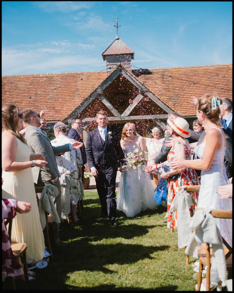 A newlywed couple walks down an outdoor aisle smiling as guests throw colorful confetti under a sunny blue sky at a Montague Farm Wedding, capturing the charm of festival-style Sussex weddings by a rustic wooden building.