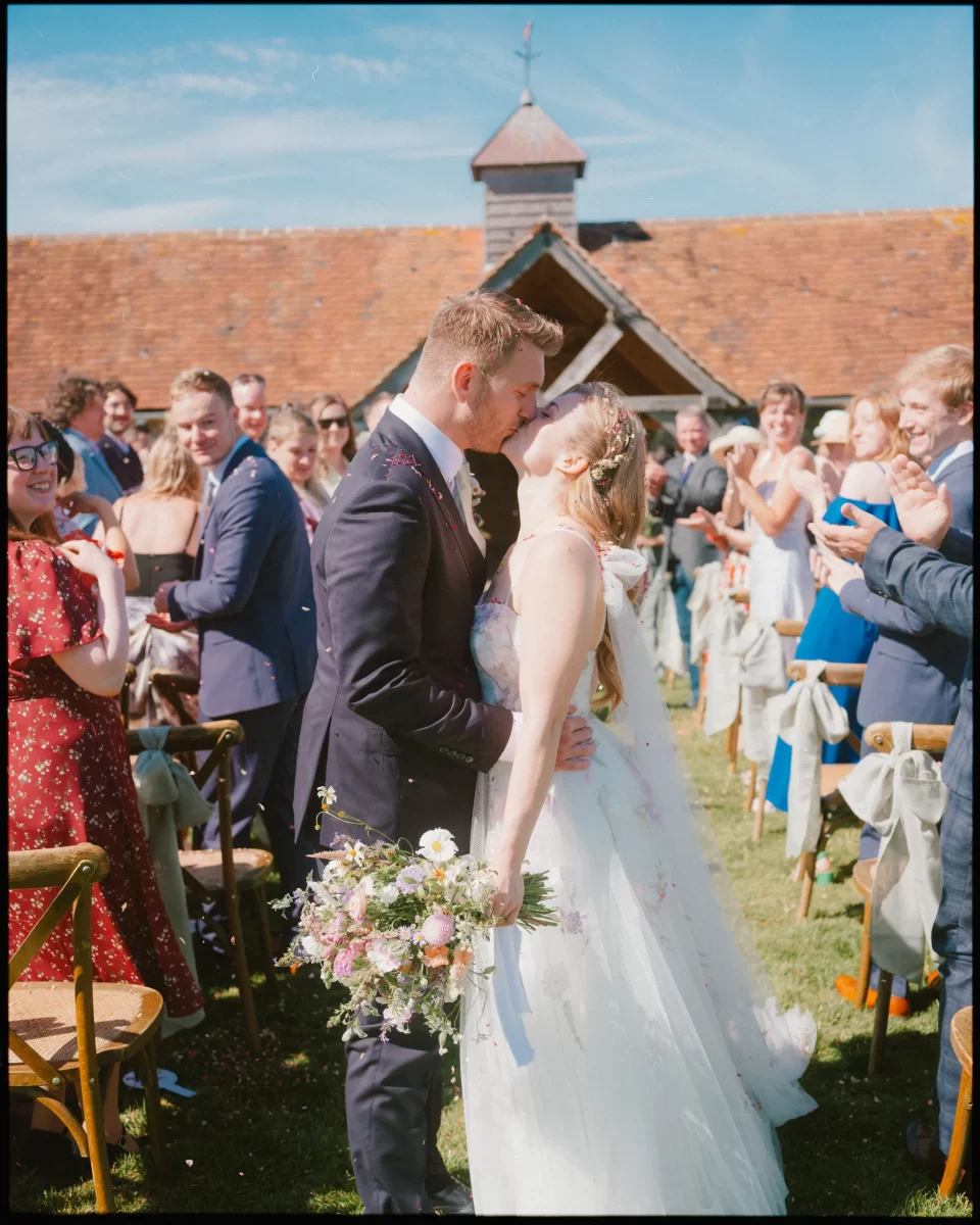 A bride and groom kiss at their Montague Farm wedding, surrounded by smiling guests who applaud and take photos. The bride holds a bouquet and wears a white dress, while the groom is in a suit—a sunny, festival-style Sussex wedding celebration.