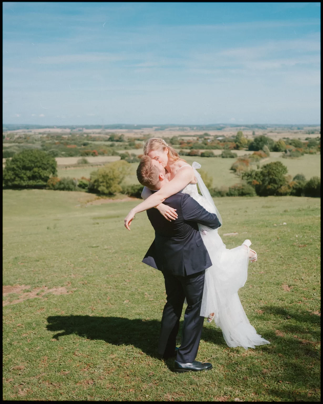 A groom in a navy suit lifts and embraces a bride in a white dress, smiling in a grassy field with scenic countryside views and blue sky—capturing the joy of a Montague Farm Wedding and festival-style Sussex weddings.