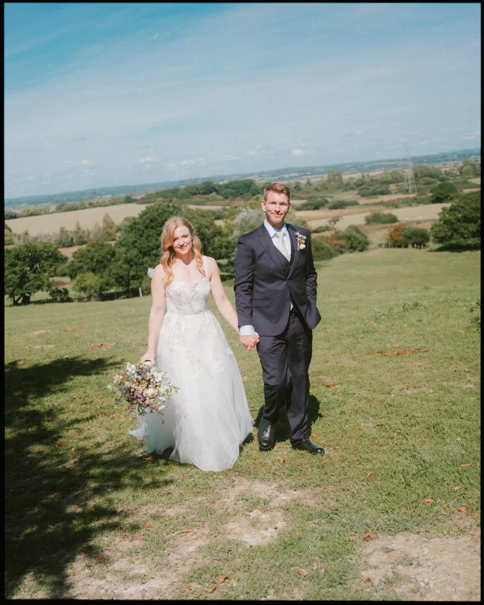 A bride in a white dress holding a bouquet and a groom in a dark suit walk hand in hand across a grassy field at Montague Farm, with trees and scenic countryside—perfect for festival-style Sussex weddings under the blue sky.