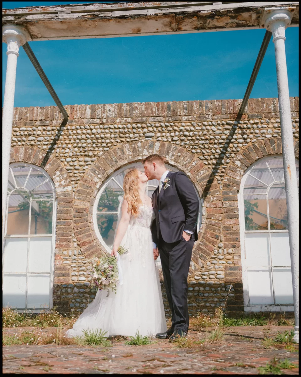 A bride in a white dress and groom in a dark suit kiss under a rustic archway at Montague Farm, celebrating their festival-style Sussex wedding in front of a stone building with a large circular window.
