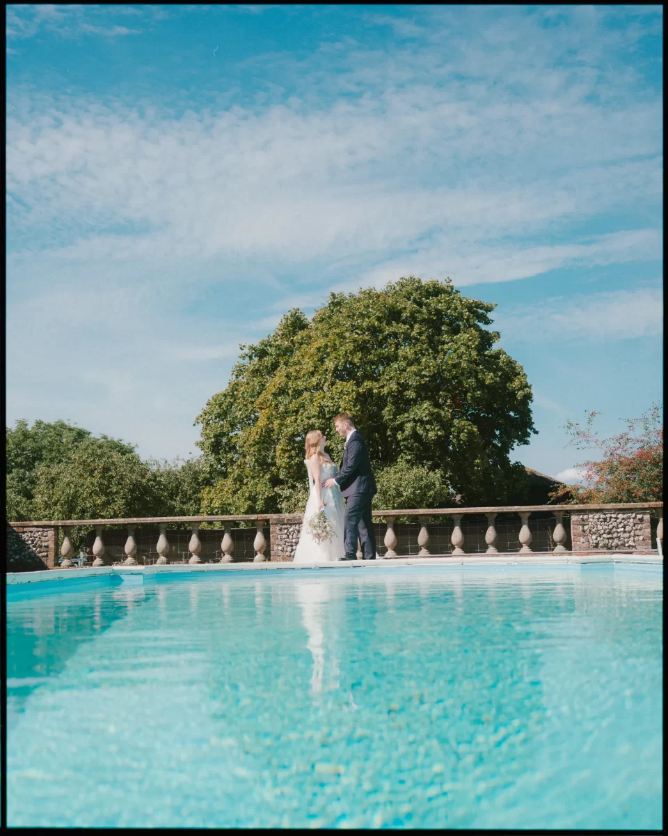 A bride and groom stand together near a bright blue swimming pool at Montague Farm, with lush green trees and a clear sky in the background, creating a peaceful and romantic scene perfect for festival-style Sussex weddings.