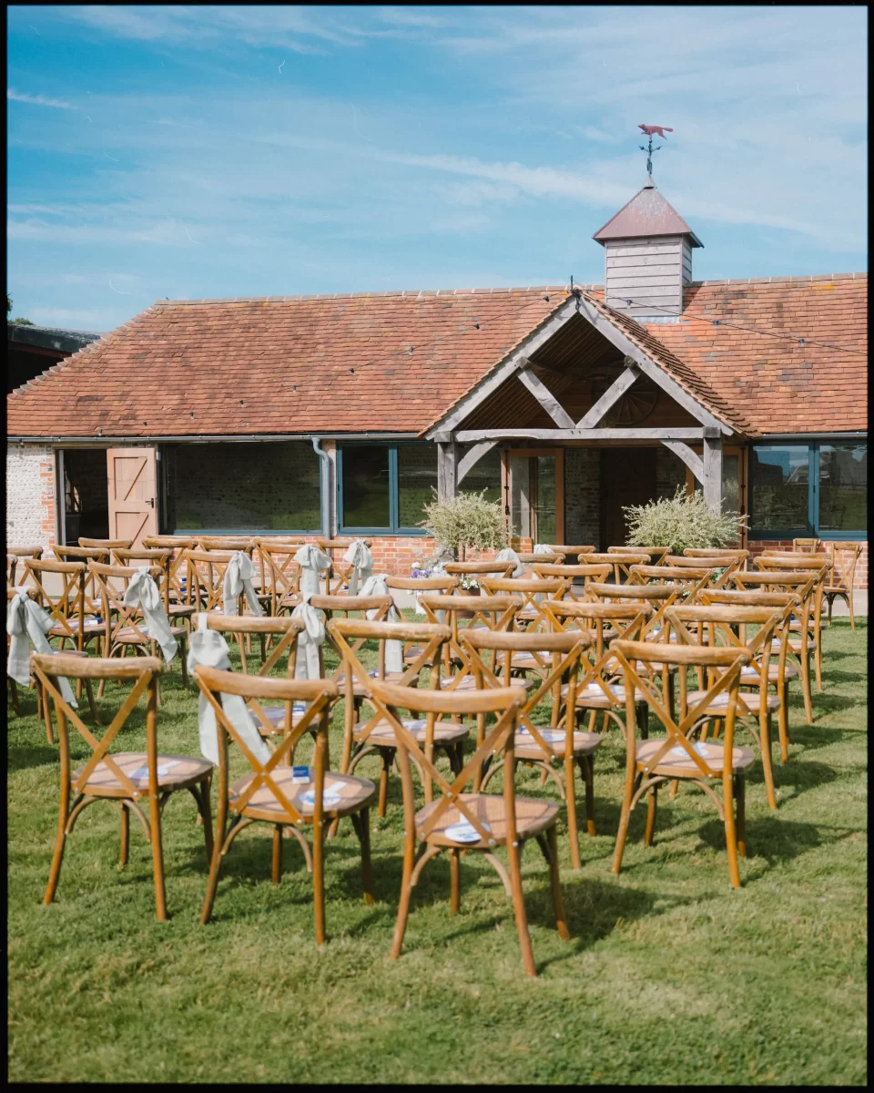 Wooden chairs arranged in rows on green grass face a rustic barn with a tiled roof and wooden porch—perfect for a Montague Farm Wedding or festival-style Sussex weddings under a clear blue sky.