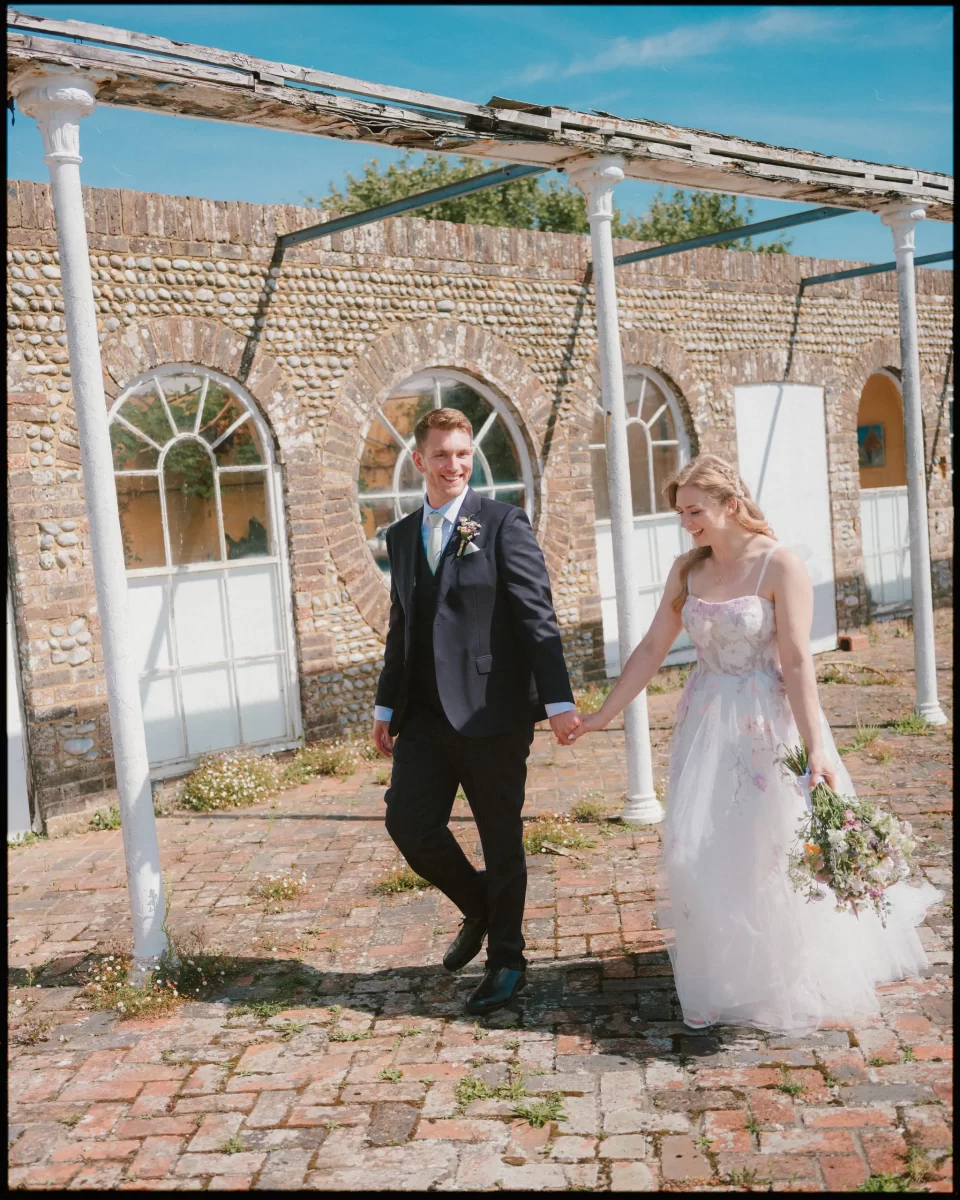 A bride and groom walk hand in hand outdoors on a sunny day at Montague Farm, smiling at each other. The bride’s white dress and bouquet shine at this festival-style Sussex wedding, framed by a rustic stone building with arched windows.