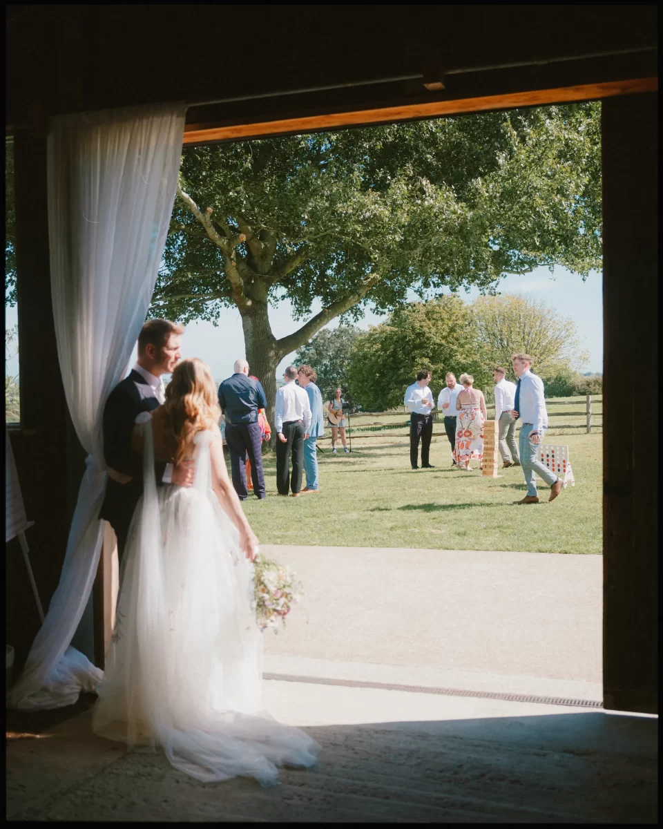 A bride and groom stand in a doorway, gazing at guests mingling on a sunny lawn beneath a large tree at their Montague Farm wedding, capturing the relaxed charm of festival-style Sussex weddings.