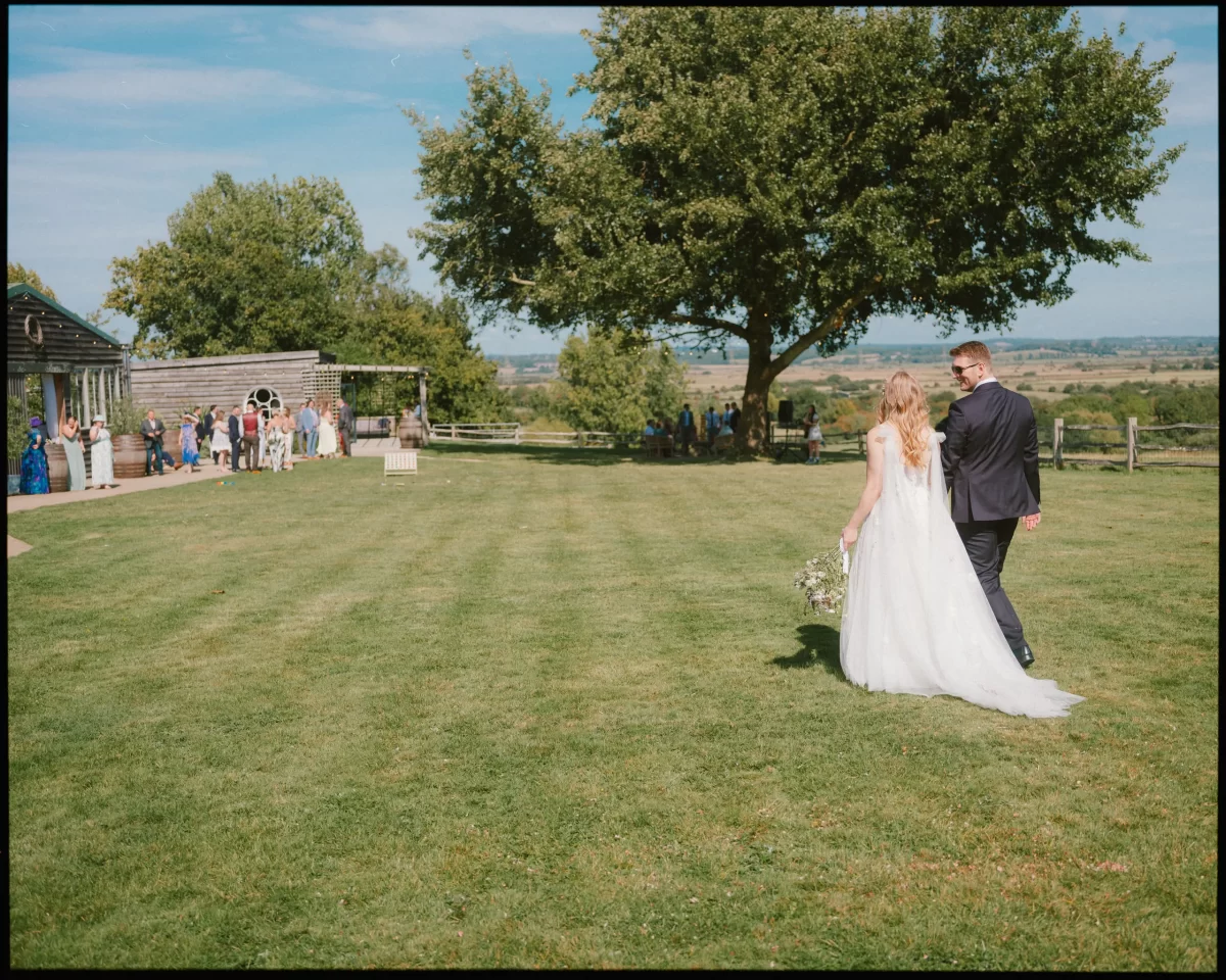 A bride and groom walk hand in hand across a large grassy lawn at Montague Farm, heading toward guests by a rustic building—perfect for festival-style Sussex weddings, with trees and open countryside in the background on a sunny day.