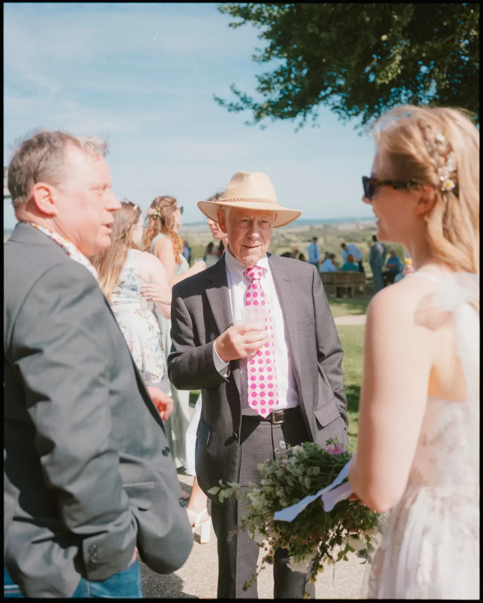 At a Montague Farm Wedding, three people chat outdoors at a festival-style Sussex wedding. A man in a straw hat and pink polka dot tie holds a drink, while another in a suit and a woman with a bouquet stand nearby amid trees and guests.