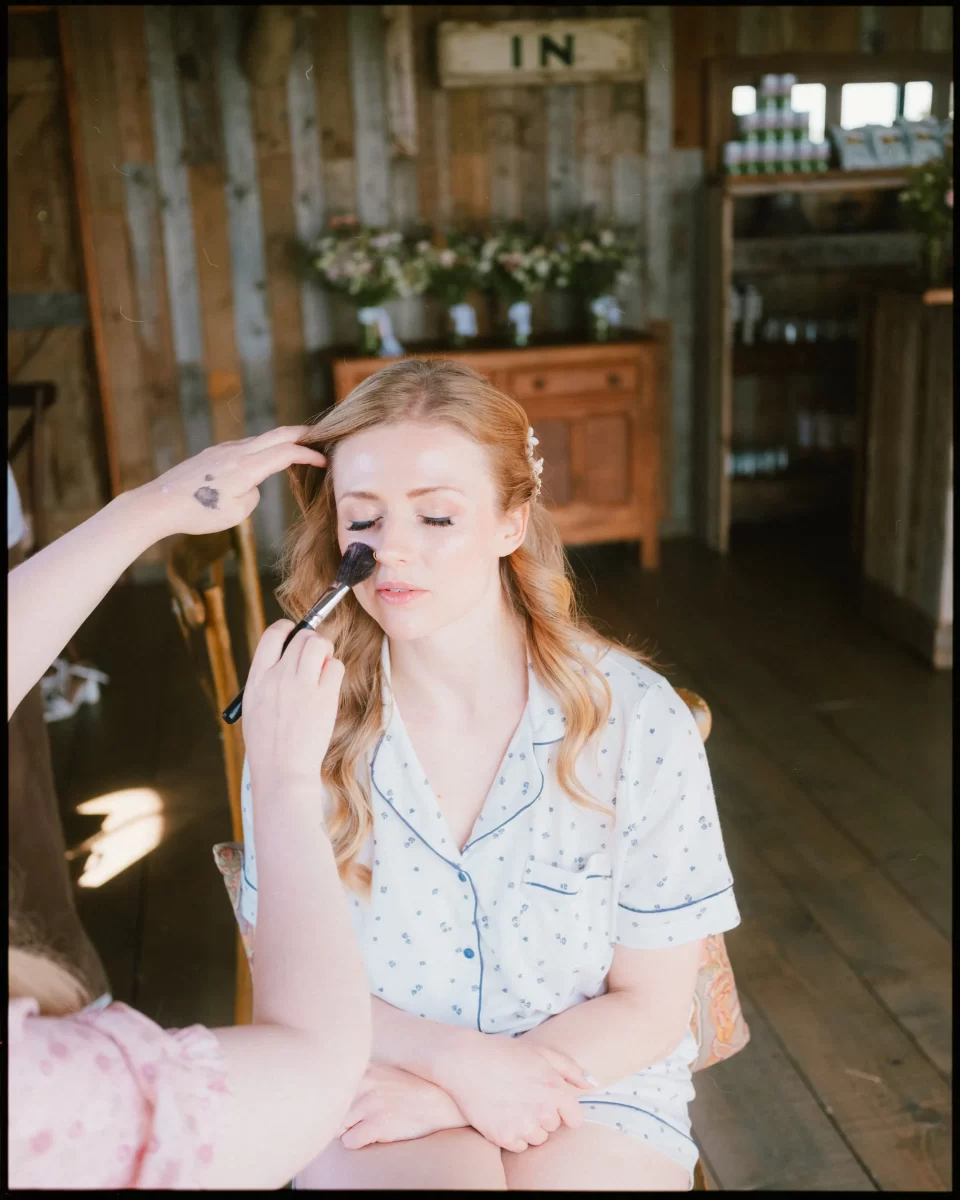 A bride getting ready in light blue pajamas sits with eyes closed while someone applies makeup to her face at a Montague Farm Wedding, set in a rustic, sunlit room with wooden walls and flower arrangements—a perfect scene for festival-style Sussex weddings.