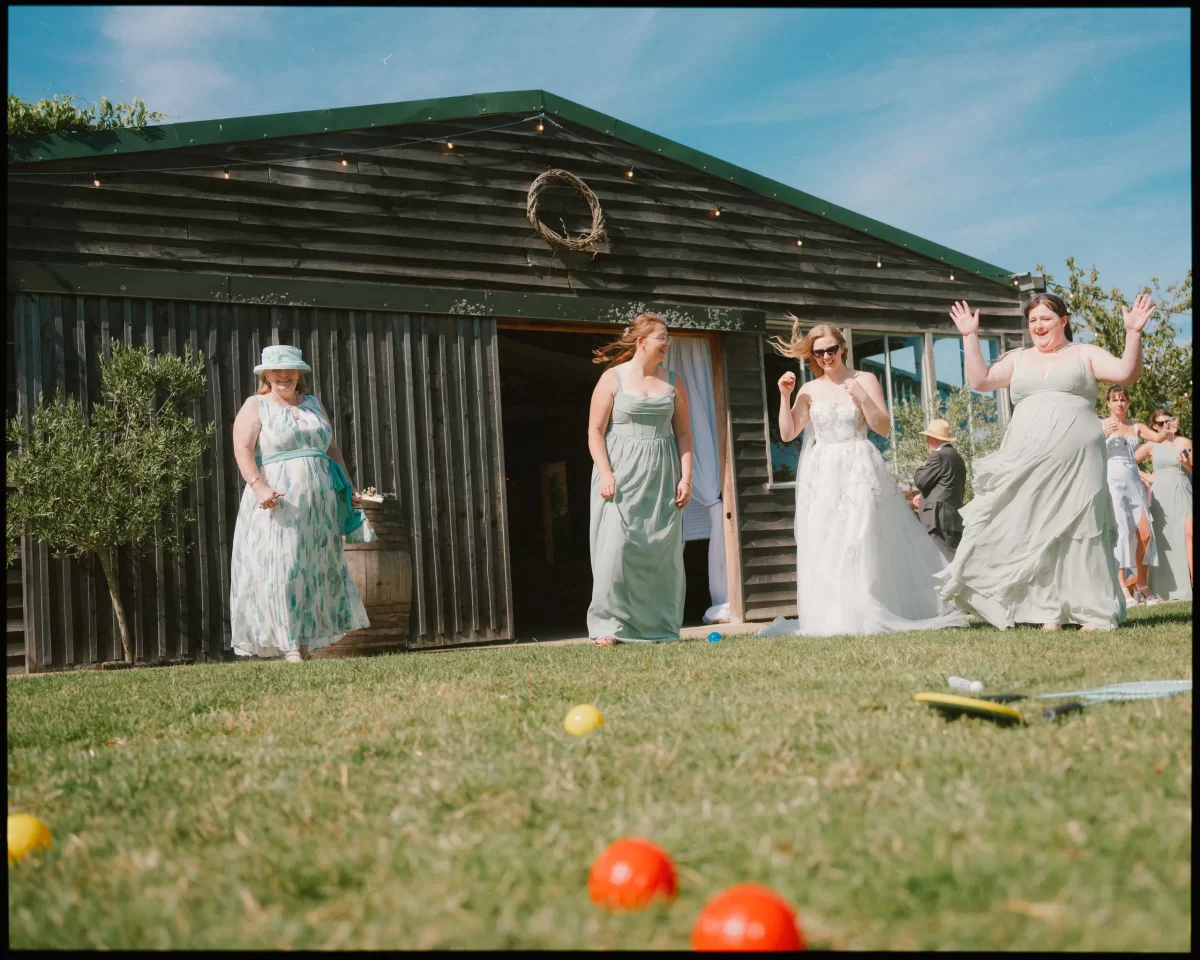 Four women in pastel dresses play lawn games on the grass outside a rustic barn at a Montague Farm Wedding. Colorful balls are scattered about, and the women laugh, embracing the joy of festival-style Sussex weddings on a sunny day.