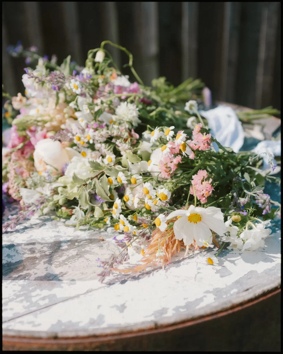 A bouquet of wildflowers, including daisies and pink blooms, rests on a weathered white wooden surface outdoors—perfect for capturing the rustic charm of a Montague Farm Wedding or festival-style Sussex weddings.
