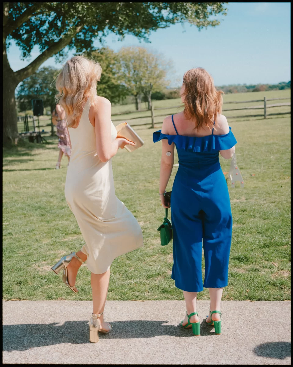 Two women stand outdoors on a sunny day at a Montague Farm Wedding. One wears a cream dress with gold heels, lifting one foot playfully, while the other dons a blue jumpsuit. Trees and a fence set the perfect festival-style Sussex weddings backdrop.