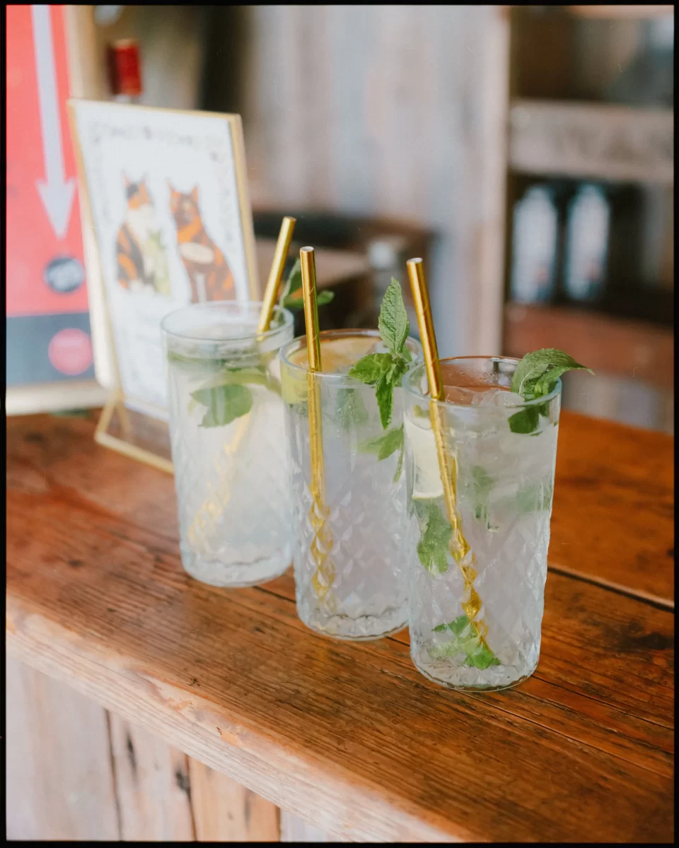 Three tall glasses of mojito cocktails with mint leaves, ice, and gold straws are lined up on a rustic wooden table—perfect for a Montague Farm Wedding or festival-style Sussex weddings in a charming café setting.