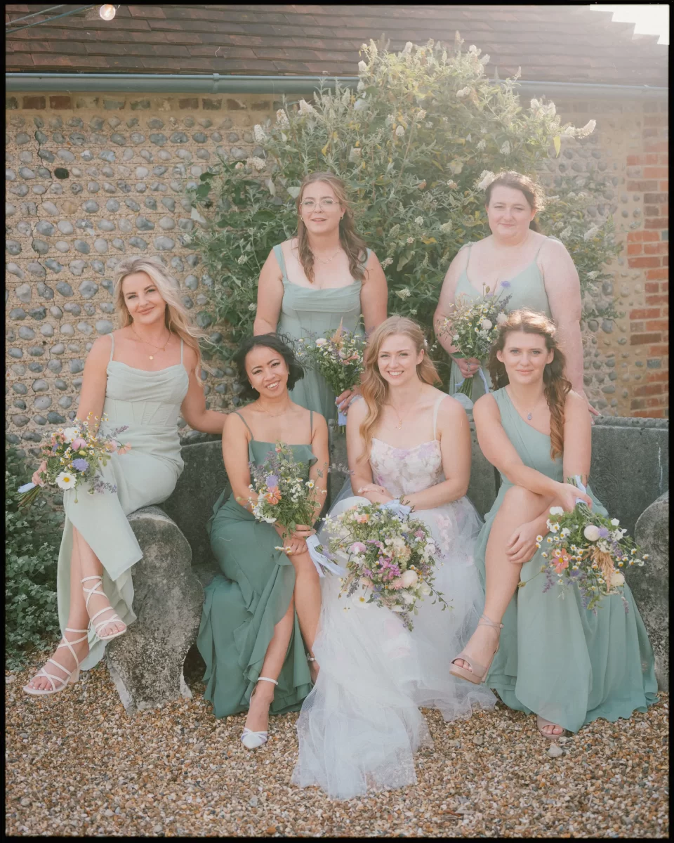 bridesmaids in pastel green and white dresses sit and stand outdoors, holding bouquets and smiling at a Montague Farm Wedding, with a stone wall and greenery in the background on a sunny day—a perfect festival-style Sussex celebration.