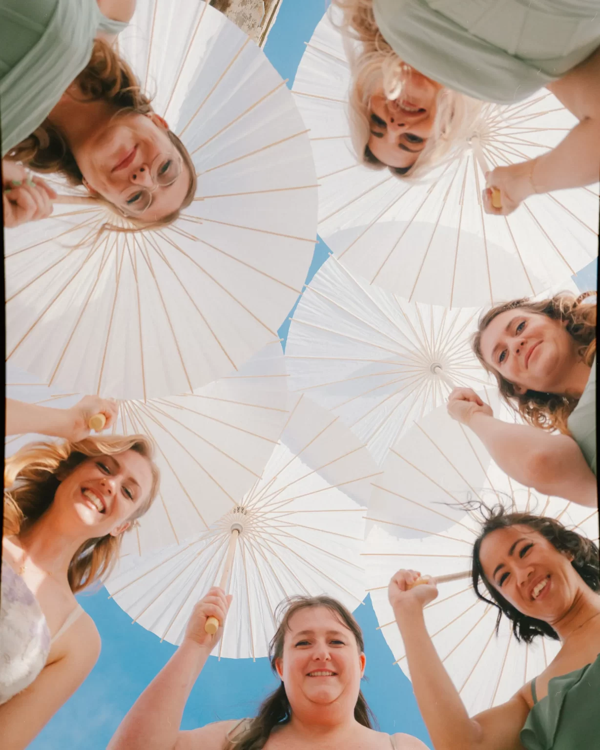 A group of six smiling women in pastel dresses stand in a circle, holding white paper parasols above their heads, looking down at the camera against a bright blue sky—a perfect moment from a Montague Farm Wedding Festival-Style Sussex wedding.
