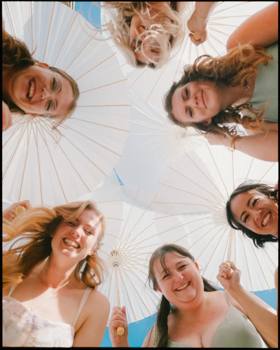 Six women in light dresses smile and hold white parasols, standing in a circle and looking down at the camera against a bright blue sky—a joyful moment at a Montague Farm Wedding, perfect for festival-style Sussex weddings.
