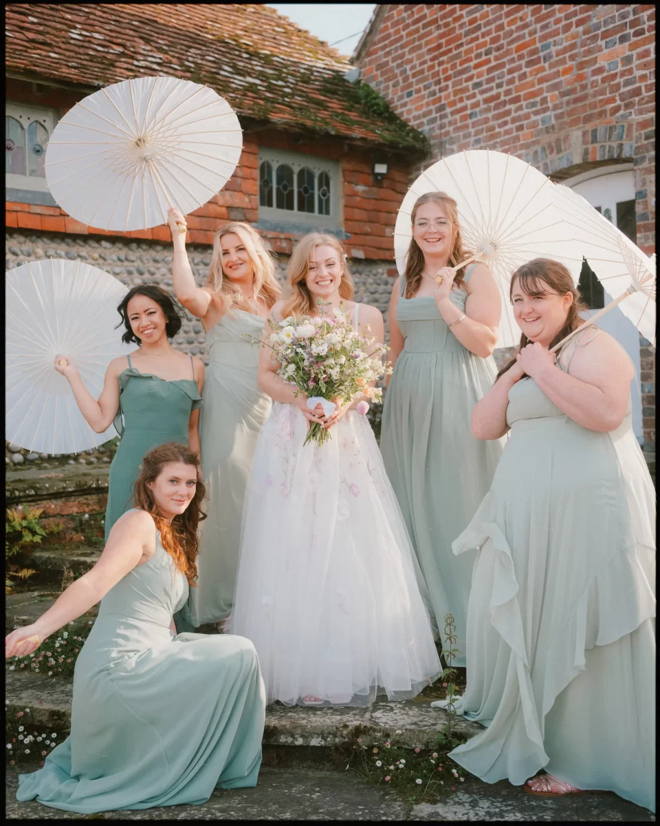 A bride in a white gown holding a bouquet stands smiling with five bridesmaids in light green dresses at a Montague Farm Wedding. Some bridesmaids hold white parasols, outdoors by brick buildings and greenery—a true festival-style Sussex wedding.
