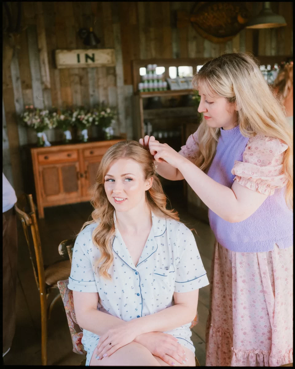 A bride in light blue pajamas sits on a chair while another styles her hair at a Montague Farm Wedding. The rustic setting, with wooden walls, flowers, and a cabinet, creates a cozy festival-style Sussex weddings atmosphere as she smiles gently at the camera.